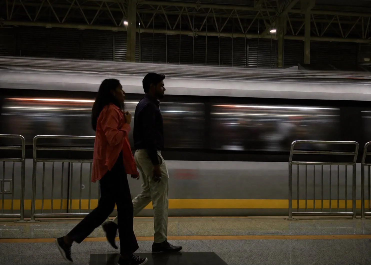 Two people walk along a platform as a metro train passes behind them.